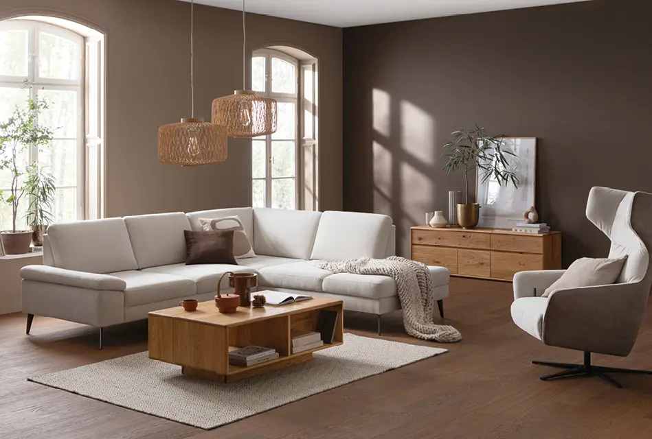 Modernes Wohnzimmer mit hellem Ecksofa, Couchtisch aus Holz und Sideboard. Warme Wandfarbe, große Fenster und dekorative Leuchten schaffen eine gemütliche Einrichtung im Wohnbereich